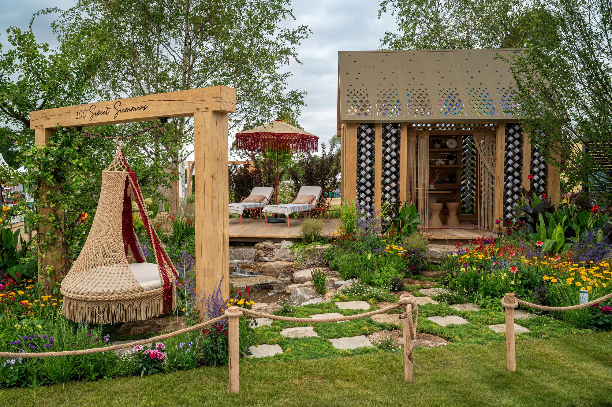 Wide view of the 100 Sweet Summers show garden at RHS Sandringham 2026 with colourful planting, water feature, macramé swing and oak garden building