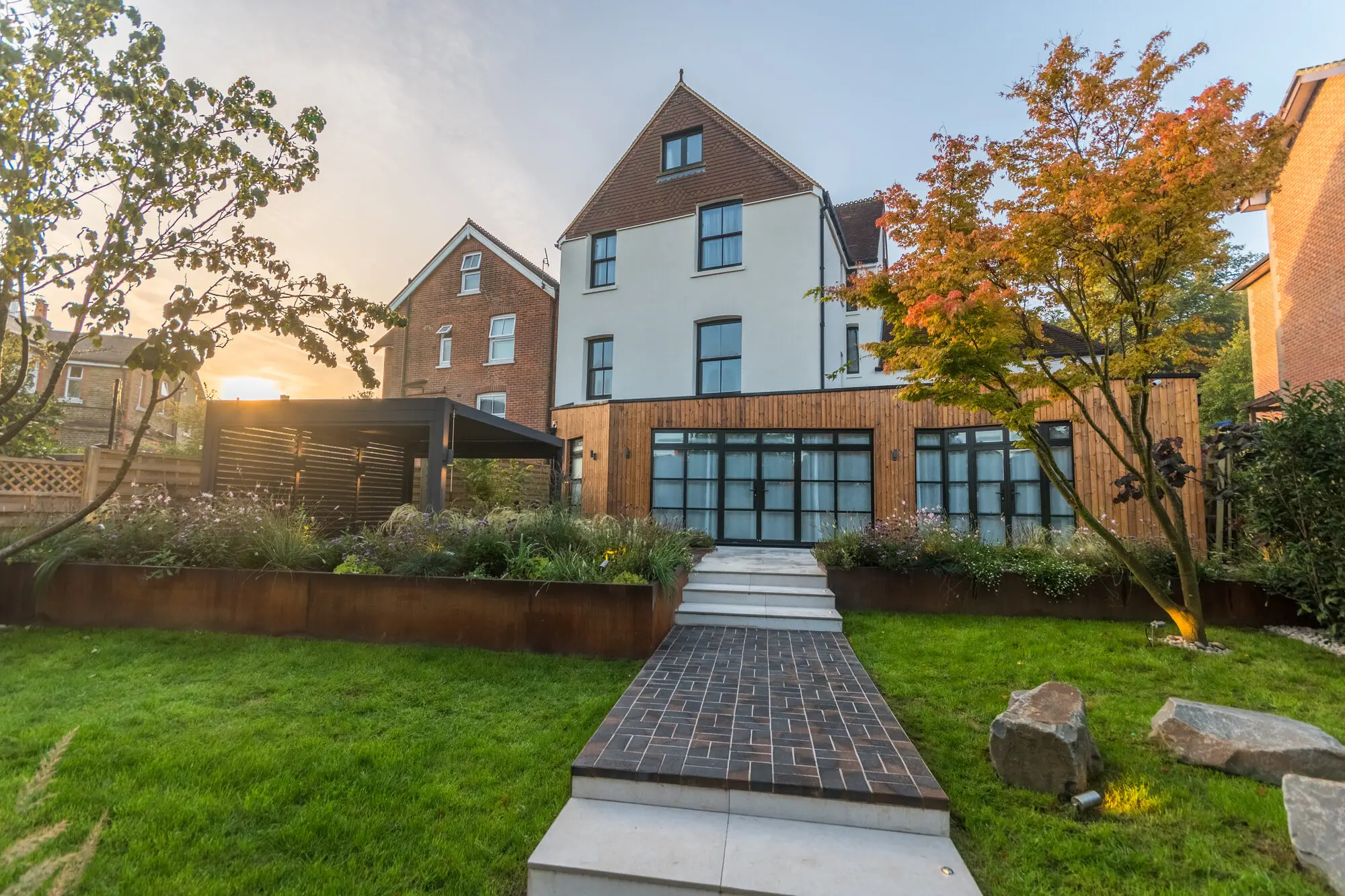 The garden seen from the lower lawn at sunrise, the brick path and stone steps rising between corten steel retaining walls to the timber-clad extension